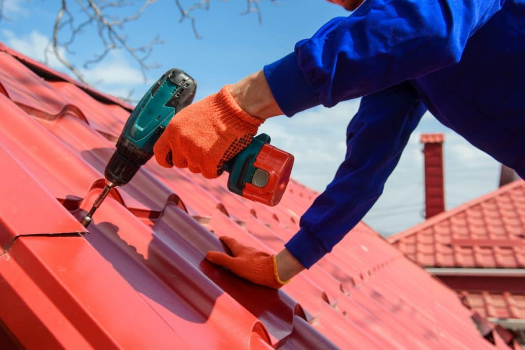 Close up of young man worker in blue overall fix a metal tile roof with screwdriver. Roofing work concept