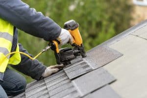 Unrecognizable roofer worker in uniform work wear using air or pneumatic nail gun and installing asphalt or bitumen tile on top of the roof under construction house