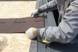 Concept of house under construction. Cropped view of professional roofer worker lays asphalt sheet on the rooftop, holding roof tile in hands
